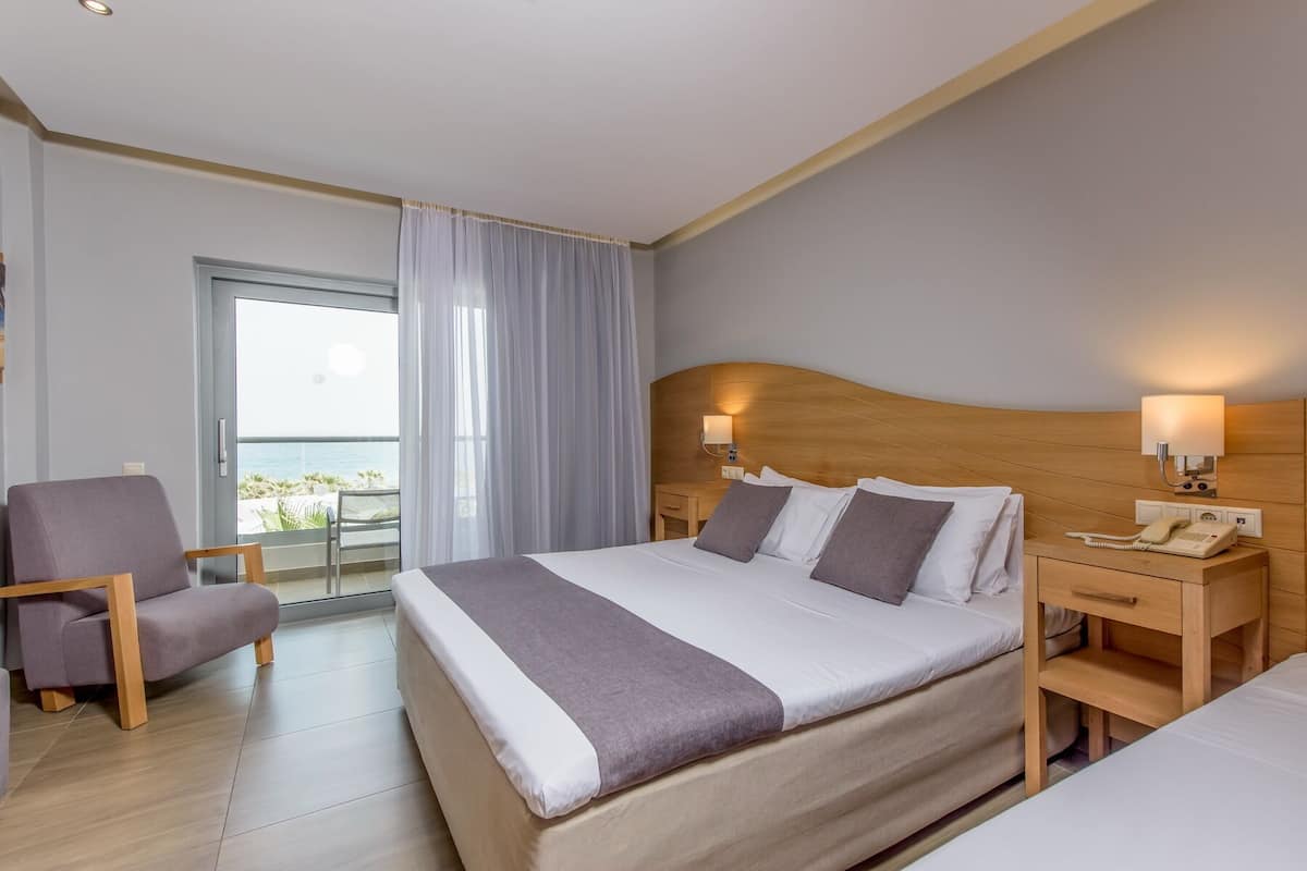 A hotel room with a large bed, a chair, a nightstand with a phone, and a view of the sea through a window.