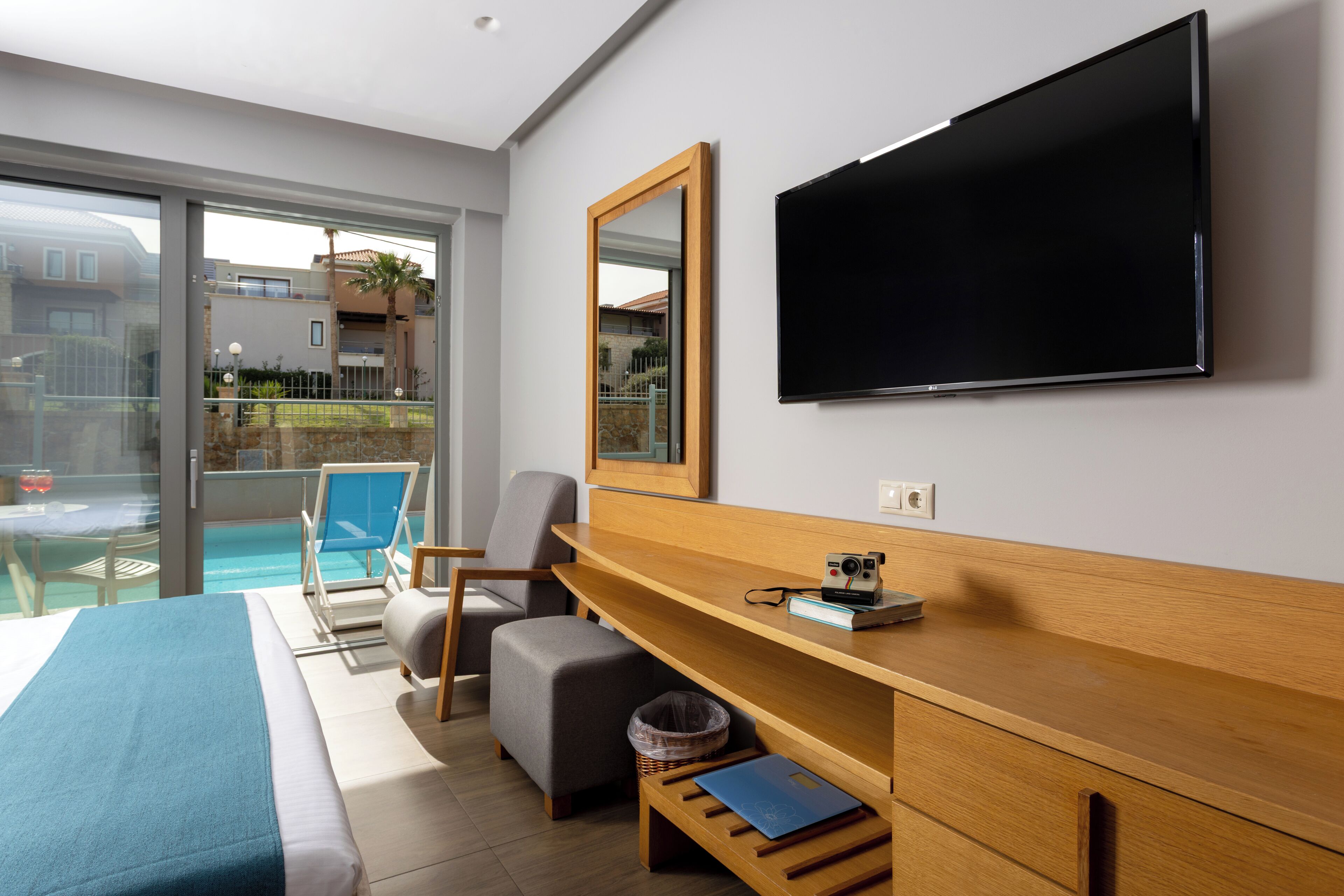 Ein modernes Hotelzimmer mit einem Bett, einem Fernseher, einem Holztisch, einem Sessel, einem kleinen Tisch und einem Balkon mit Pool.