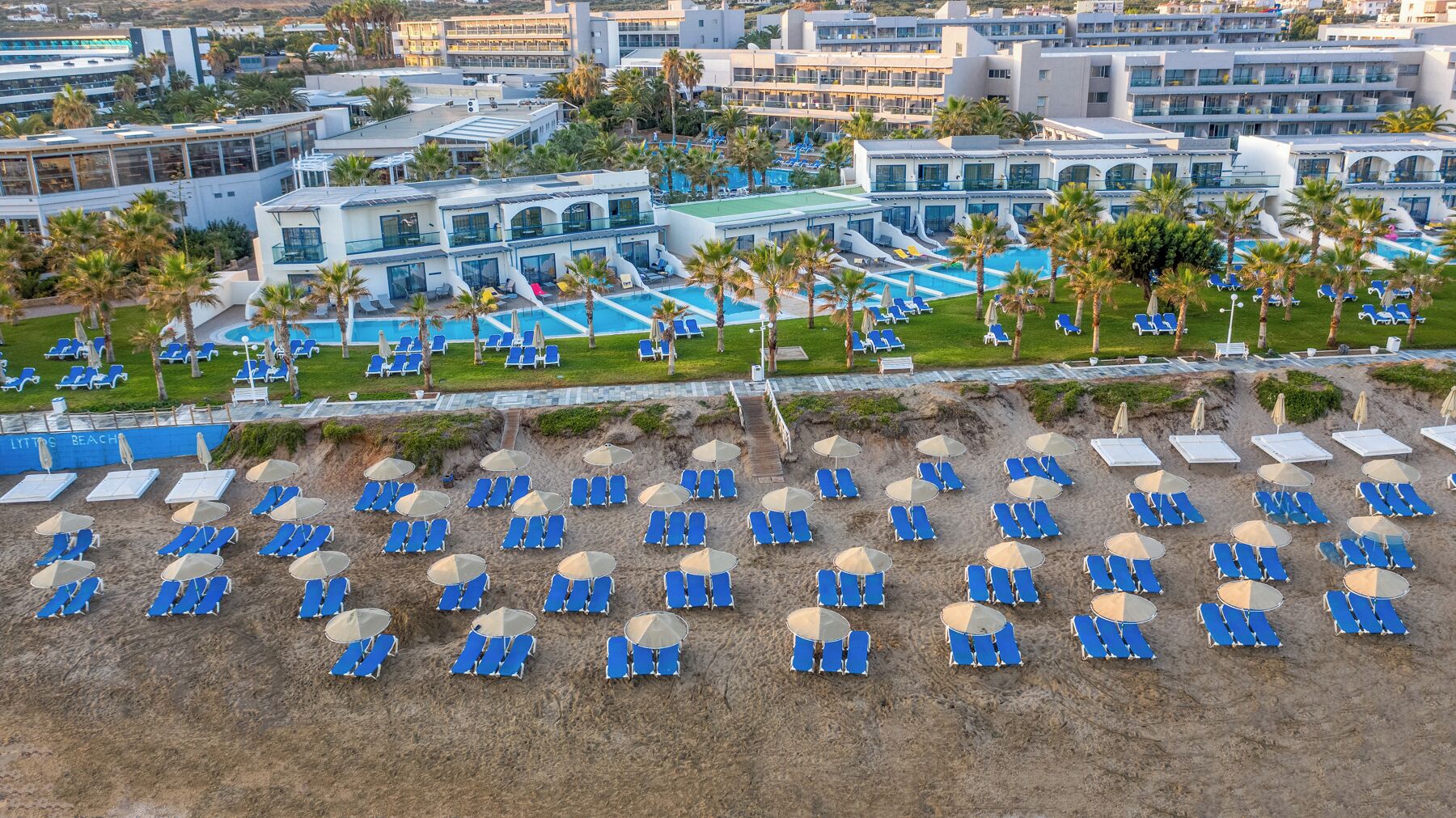 A resort with multiple swimming pools, a beach area with sun loungers and umbrellas, and modern buildings with balconies.