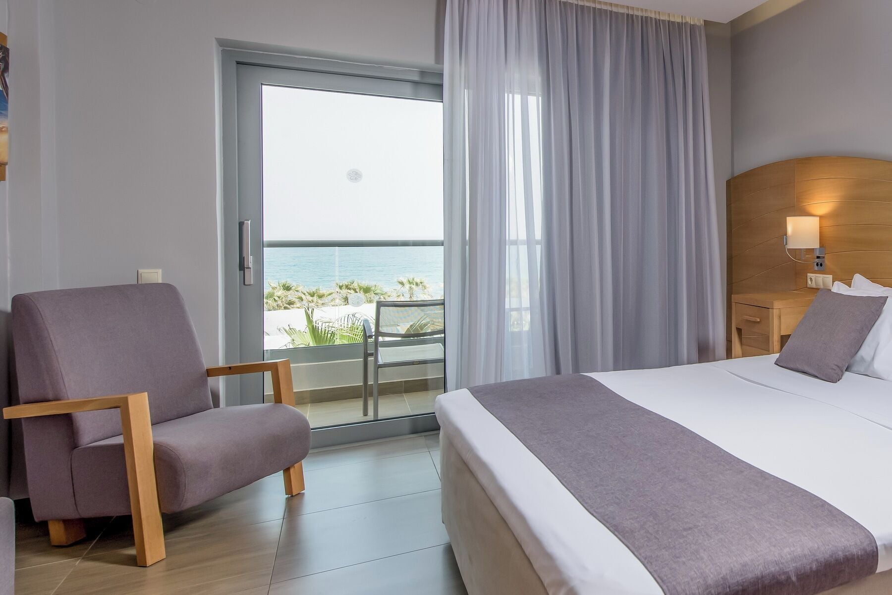 A hotel room with a bed, a chair, a nightstand, and a view of the sea through a large window.