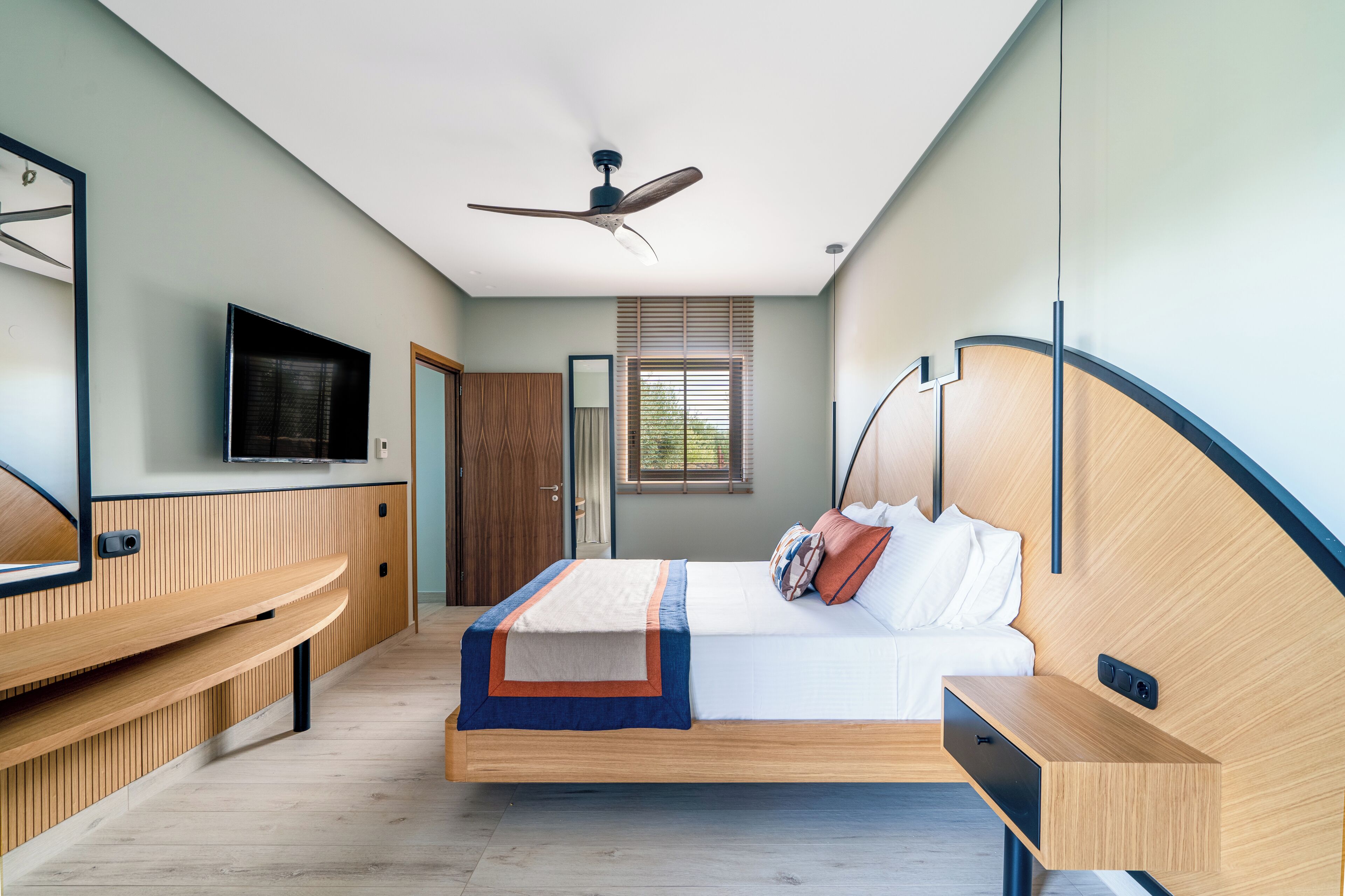 Ein modernes Schlafzimmer mit Bett, Nachttisch, integriertem Fernseher, Deckenventilator und einem Fenster mit Jalousien.