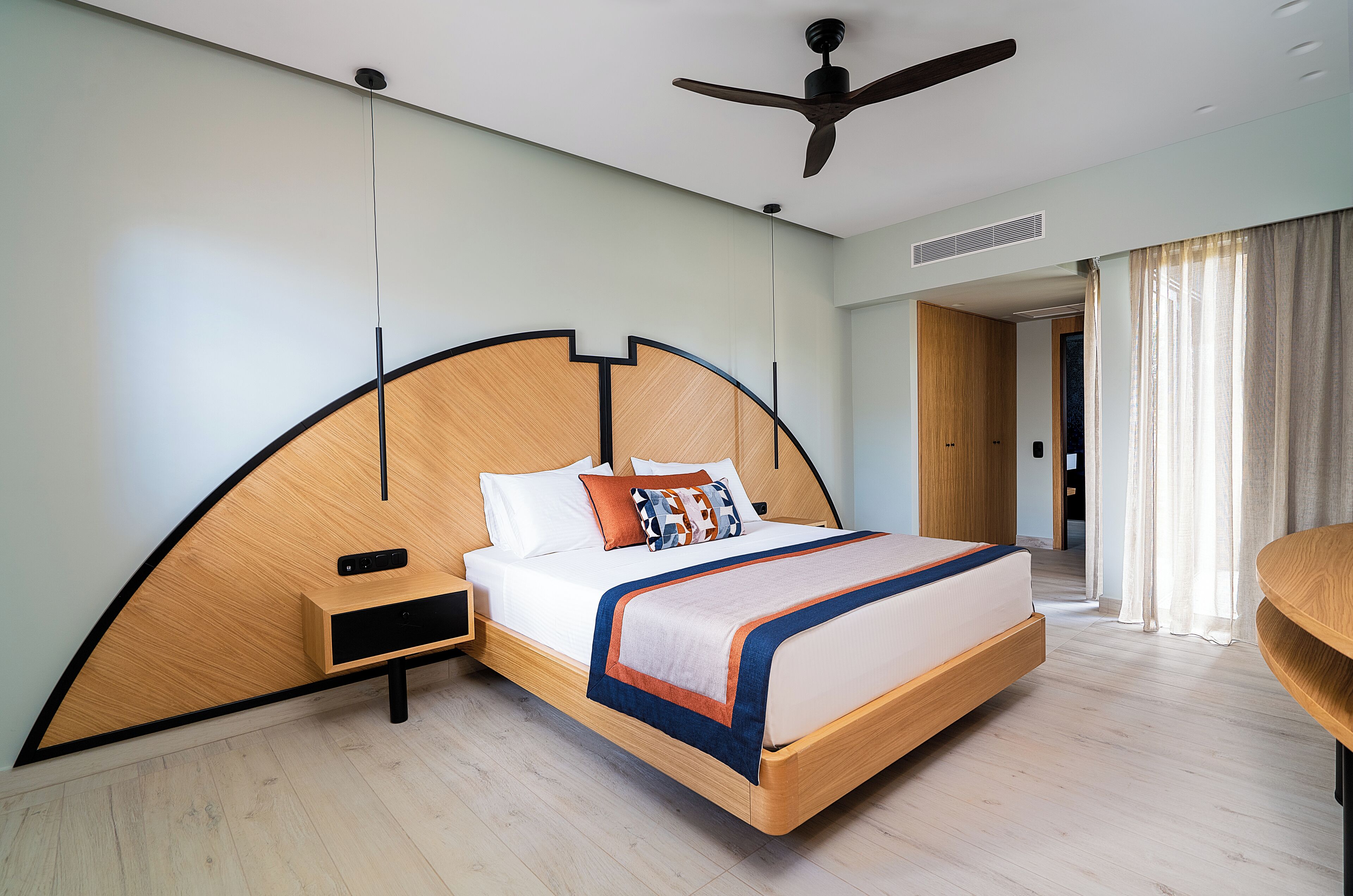 A modern hotel room with a curved headboard, a bed with a blue and orange bedspread, a nightstand, and a ceiling fan.