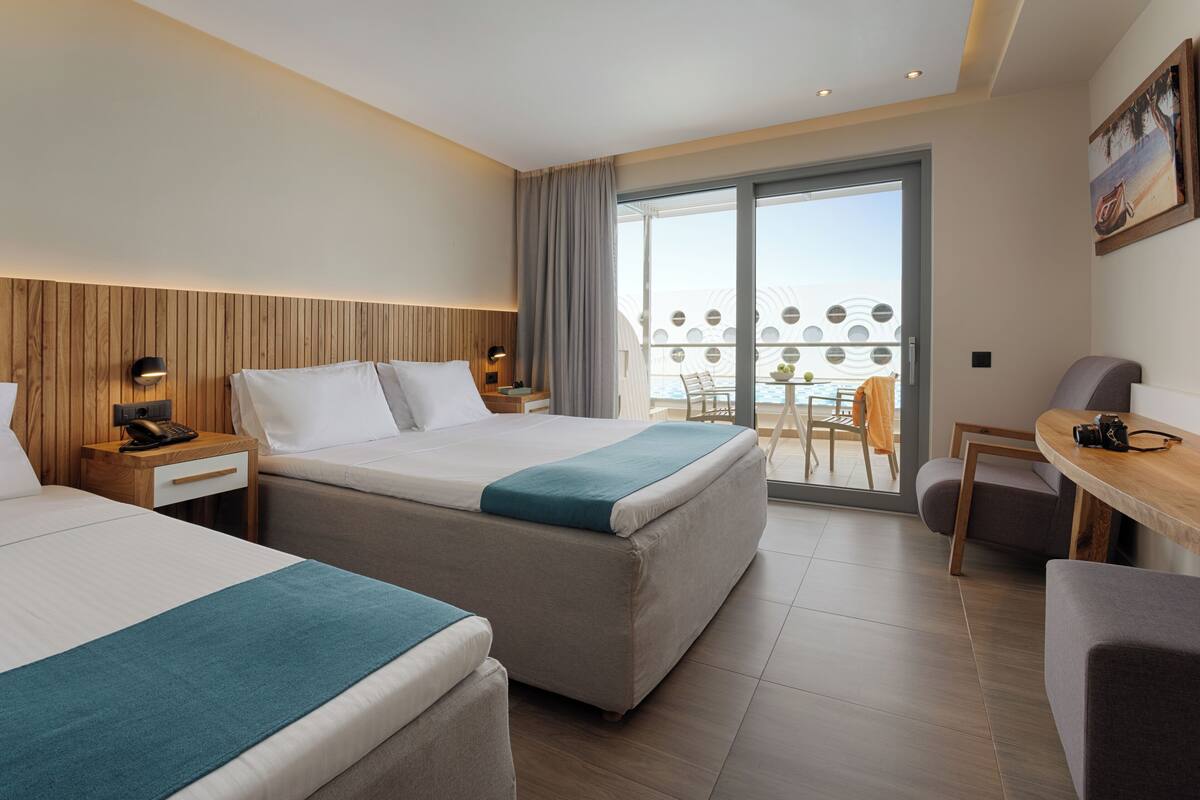 A hotel room with two beds, a desk, a chair, a balcony with a table and chairs, and a view of the sea.