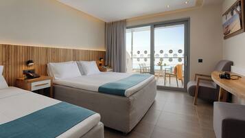 A hotel room with two beds, a desk, a chair, a balcony with a table and chairs, and a view of the sea.