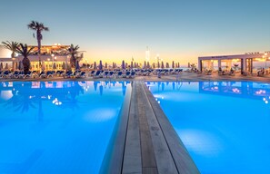 Zona de piscina con sillones de descanso y sombrillas, un edificio con balcón y vista al atardecer.