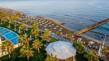 Vue aérienne d’un complexe hôtelier en bord de mer, avec une piscine, des transats, des parasols et une grande zone abritée.