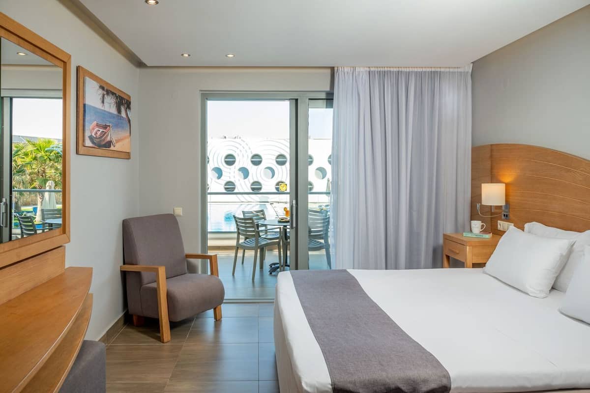 A hotel room with a bed, a chair, a desk, a lamp, and a view of an outdoor area with a table and chairs.