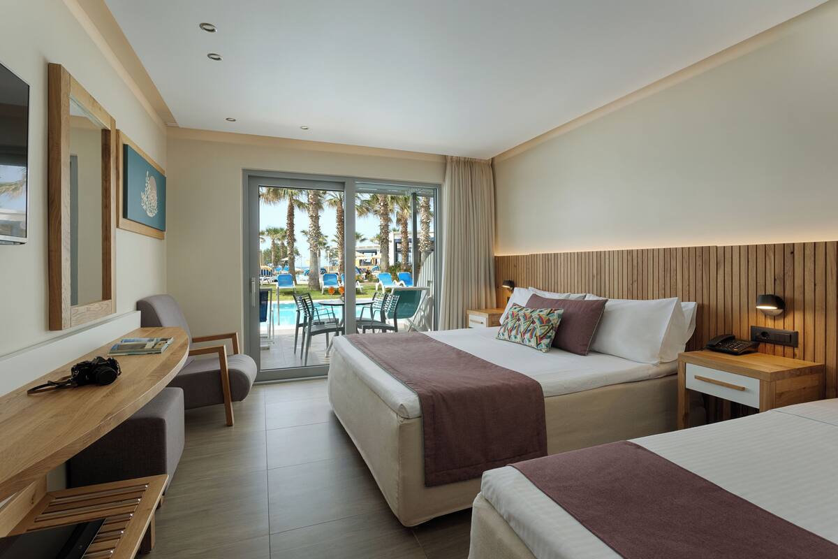 A hotel room with two beds, a desk, a chair, a small table, a nightstand, and a view of the pool area.