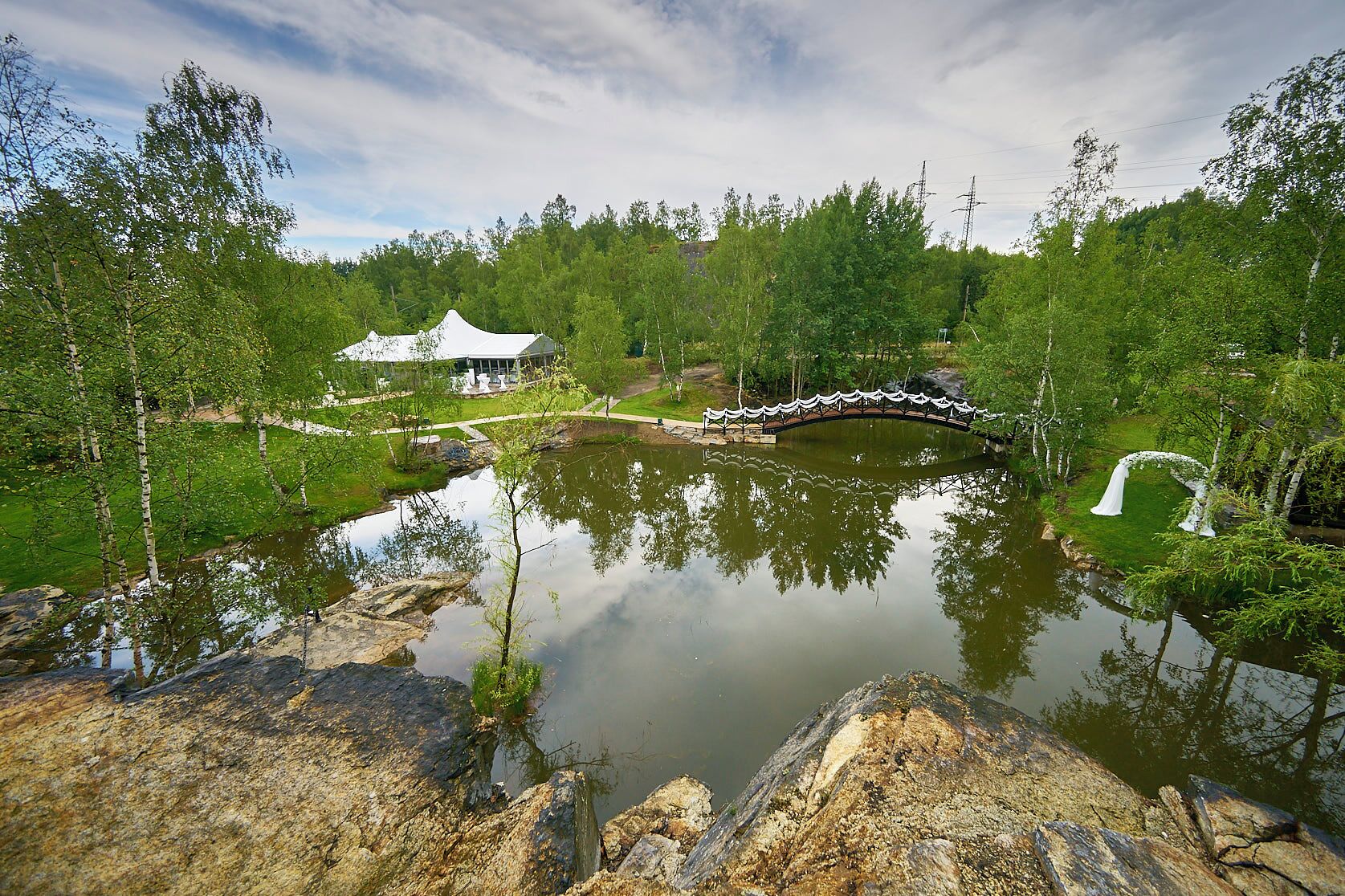 Un tranquillo paesaggio lacustre con un ponte, un gazebo e un edificio immerso nel verde.
