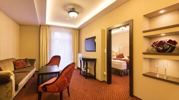 Une chambre d’hôtel avec un canapé, une table à manger et des chaises, un téléviseur, une petite table avec un vase et un miroir qui reflète un lit.