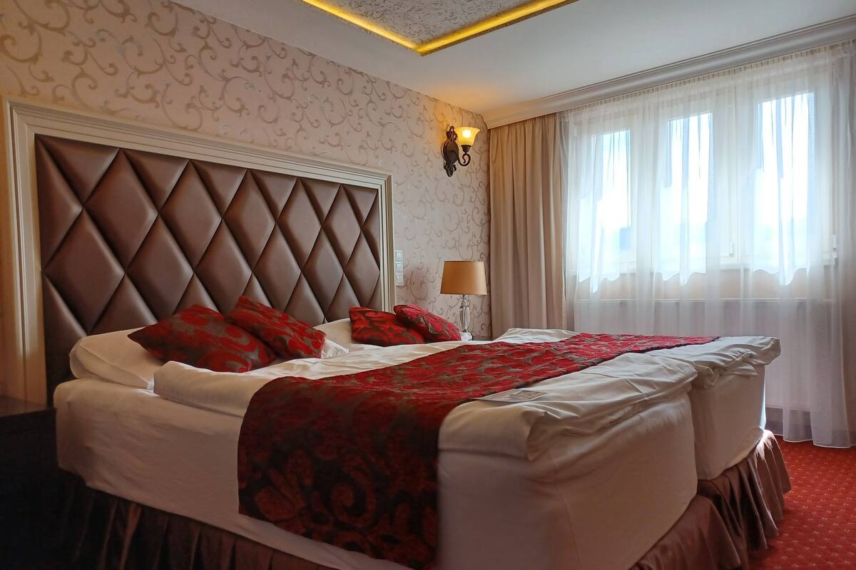 Quarto de hotel com uma cama grande, cabeceira, luminária e janela com cortinas.