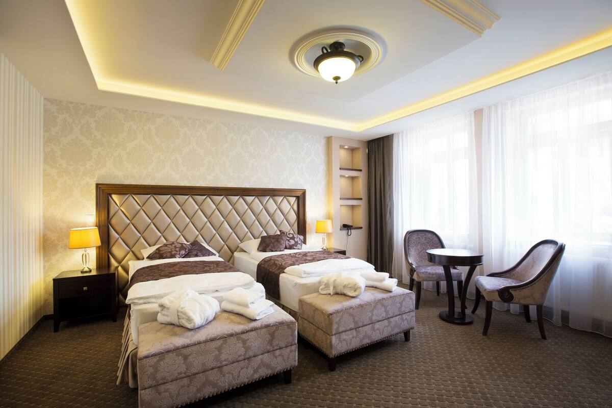 Quarto de hotel com uma cama grande, duas mesas de cabeceira, uma cadeira, uma mesinha e uma janela com cortinas.