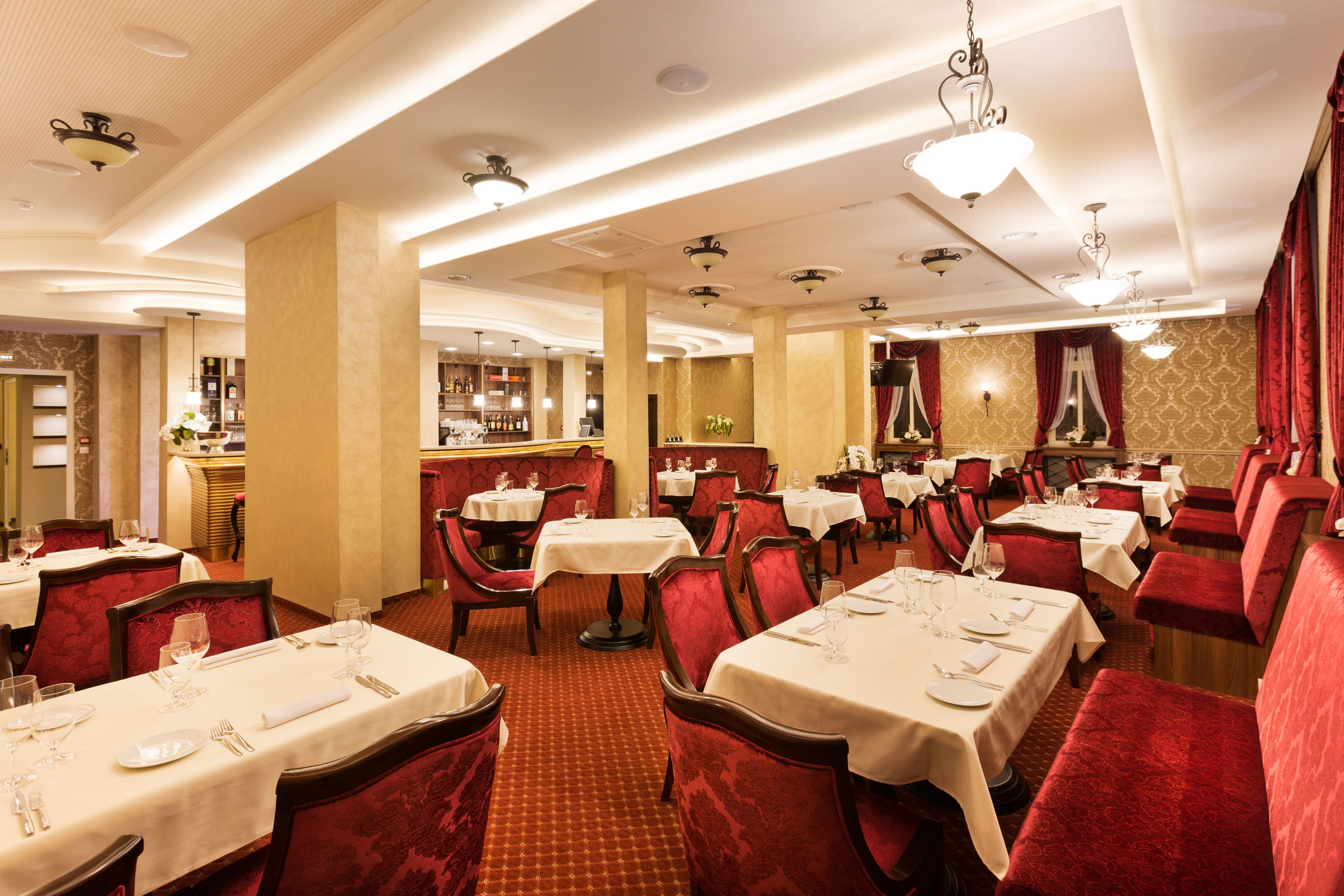 Un restaurante con varias mesas, sillas tapizadas en rojo, manteles blancos y una zona de bar al fondo.
