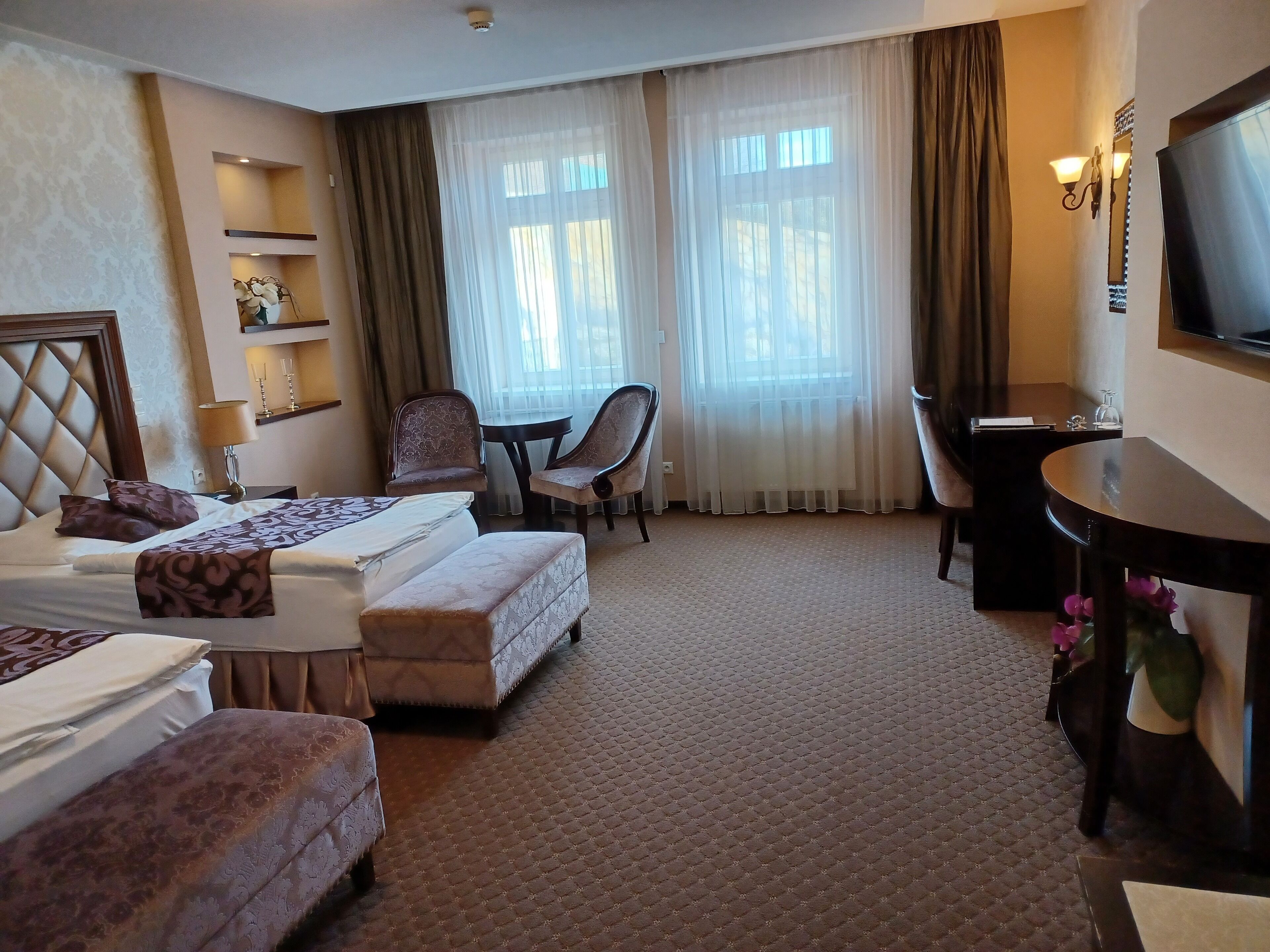 A hotel room with a bed, two chairs, a desk, a television, and a window with curtains.