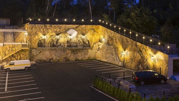 Un estacionamiento con una camioneta blanca y un coche negro, una escalera que sube a un edificio, y un gran muro de roca con un mural.