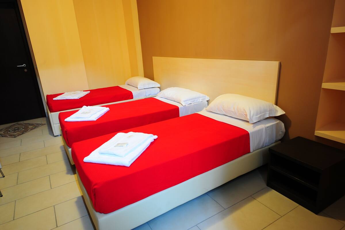 A hotel room with three single beds, each with a red bedspread and white pillows, and a wooden headboard.