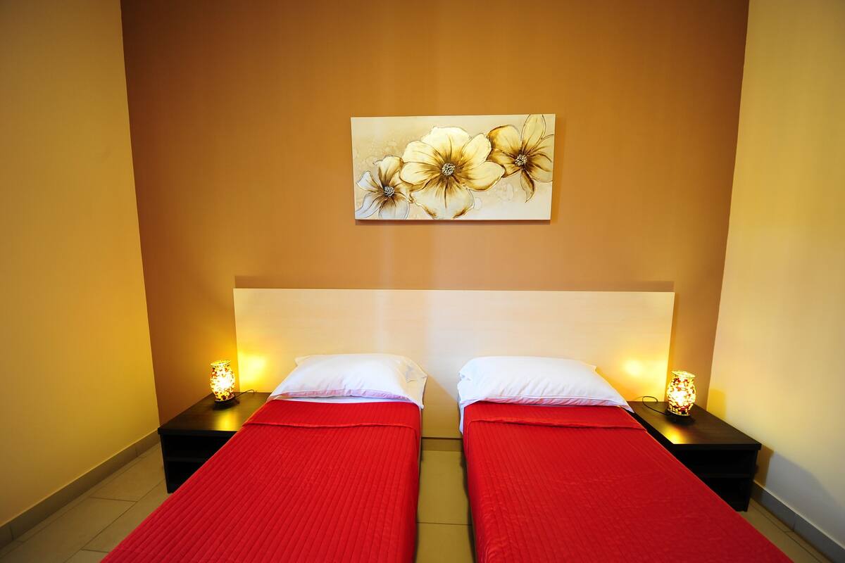 A hotel room with two beds, each with a red bedspread and white pillows, flanked by nightstands with lamps. A floral artwork is displayed on the wall above the beds.