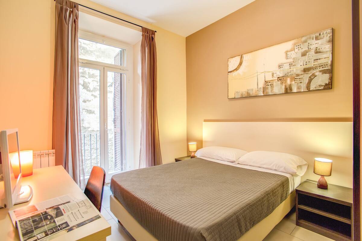 A hotel room with a large bed, a desk with a computer, a chair, a window with curtains, and a wall-mounted artwork.