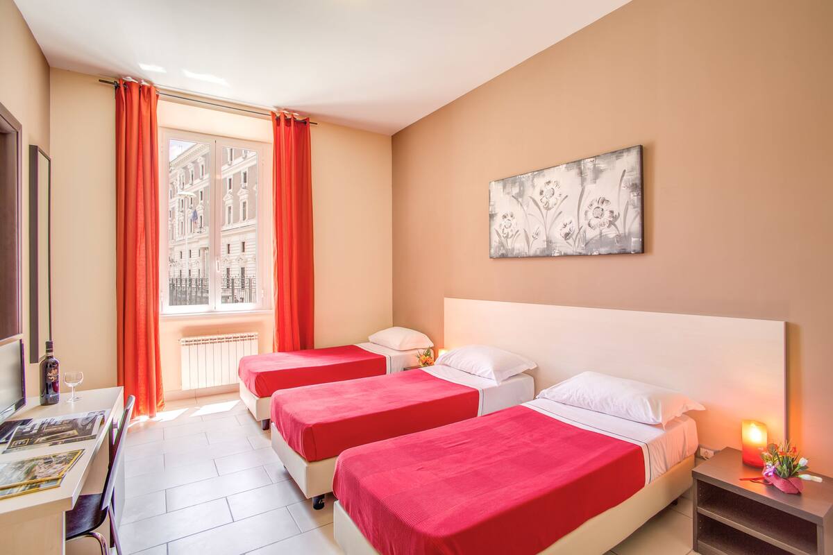 A hotel room with two beds, a desk with a chair, a nightstand with a lamp, and a window with curtains.