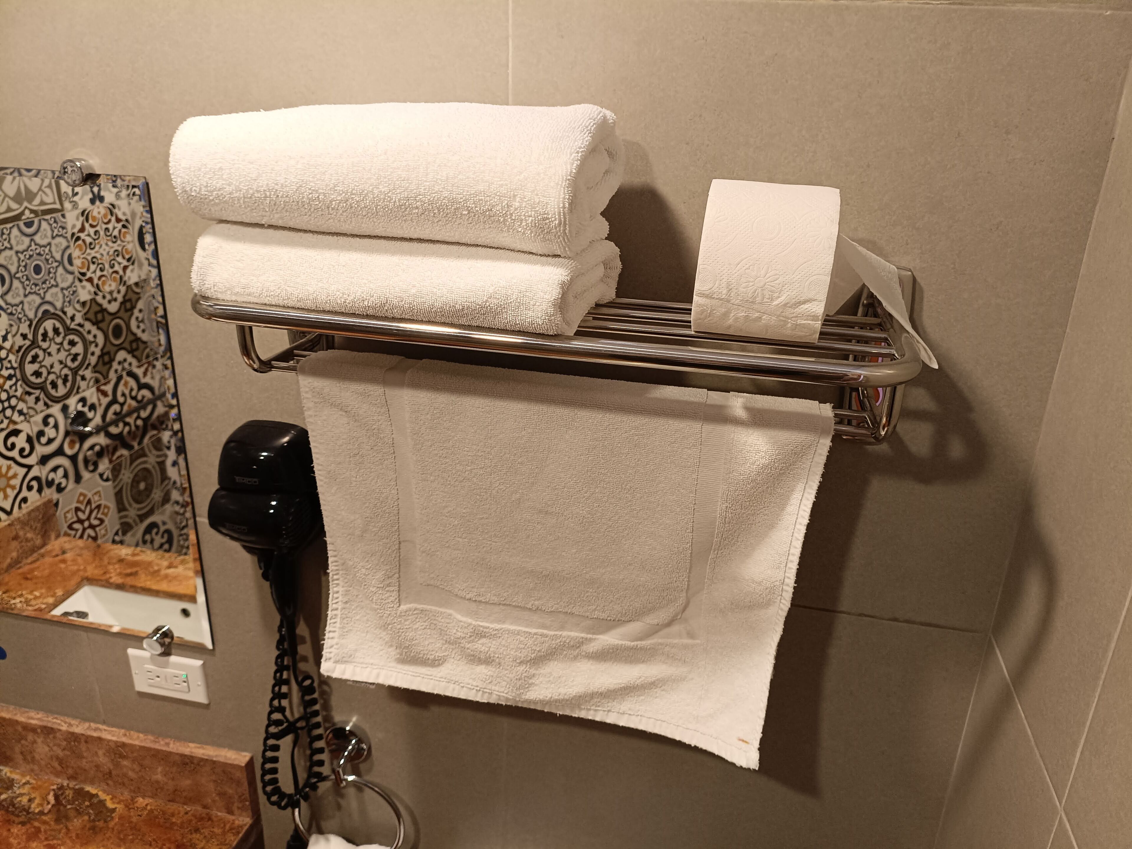 A bathroom with a towel rack holding two folded towels and a roll of toilet paper. A hairdryer is on the floor next to the towel rack.