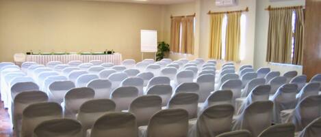 Un salón de conferencias con filas de sillas cubiertas con fundas blancas, una mesa con mantel blanco y una pizarra.