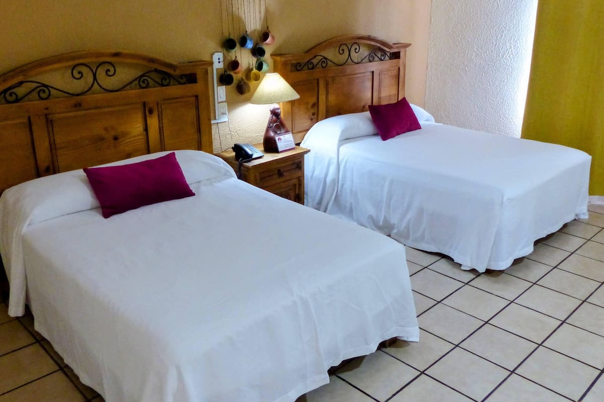 Dos camas individuales con sábanas blancas y almohadas moradas, un cabecero de madera, una mesita de noche con una lámpara y un adorno en la pared.