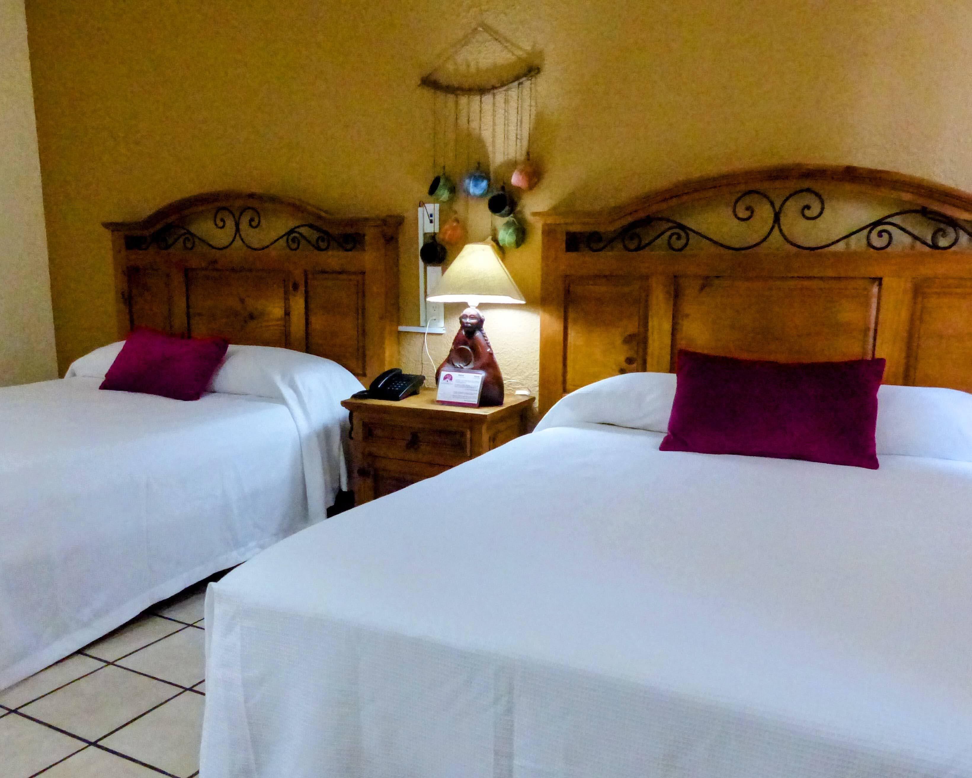 A hotel room with two single beds, each with a wooden headboard and a white bedspread. There is a nightstand between the beds with a lamp, a telephone, and a sign. The room has a wooden ceiling with a decorative hanging element.