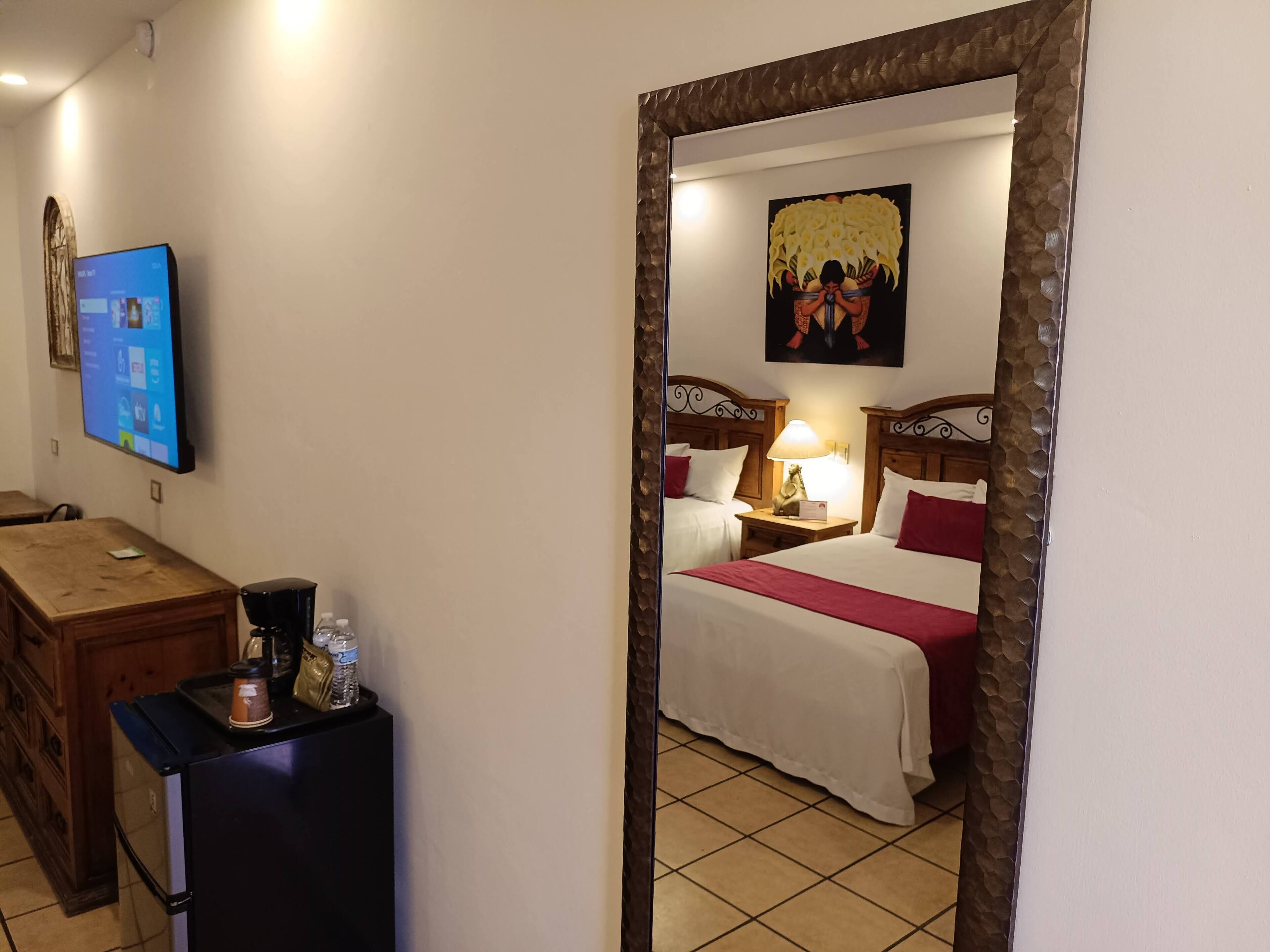 A hotel room with a bed, a desk with a computer, a coffee maker, and a mirror reflecting the room.