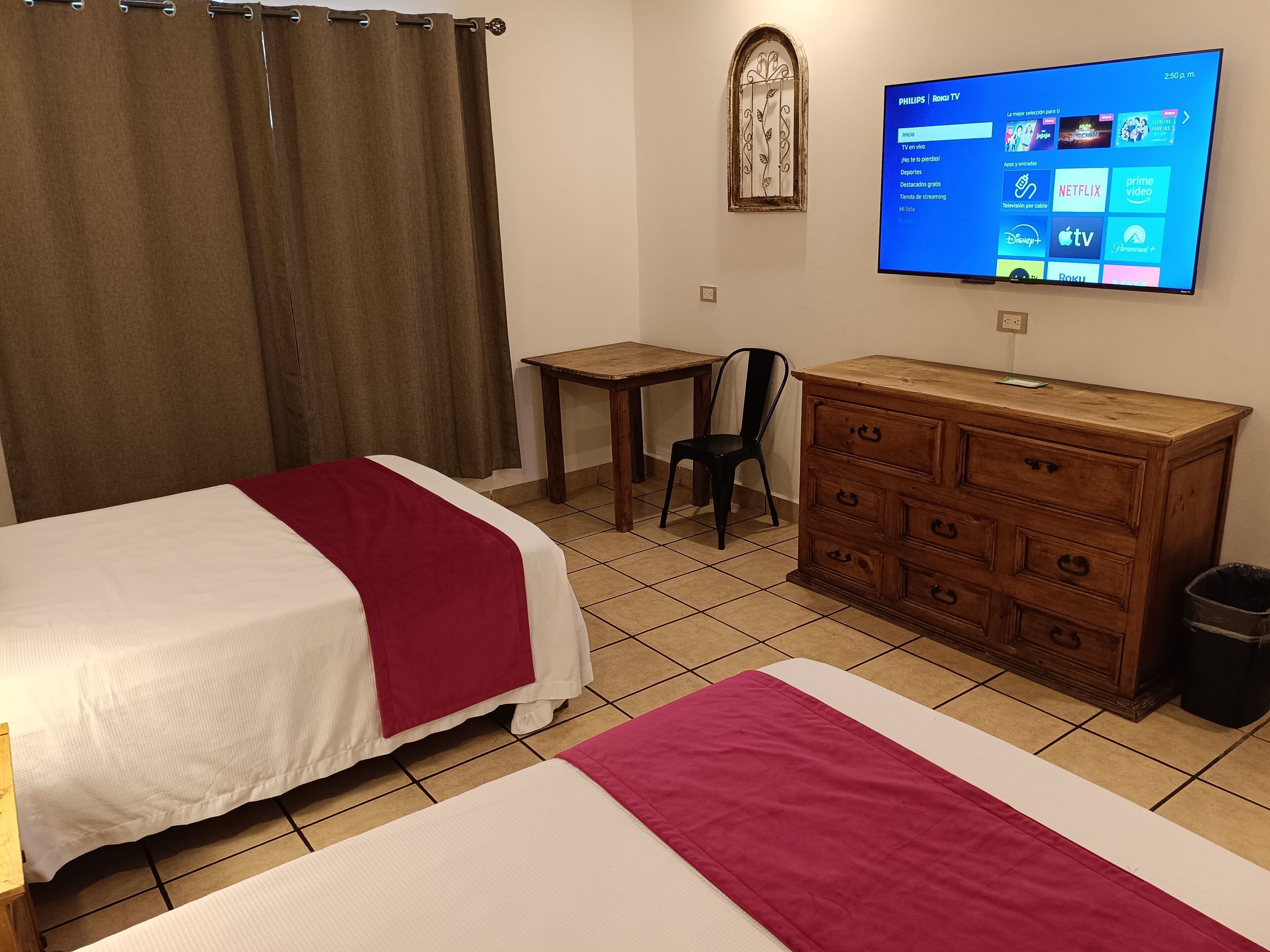 A hotel room with two beds, a wooden dresser, a desk, a chair, a television mounted on the wall, and a window with curtains.