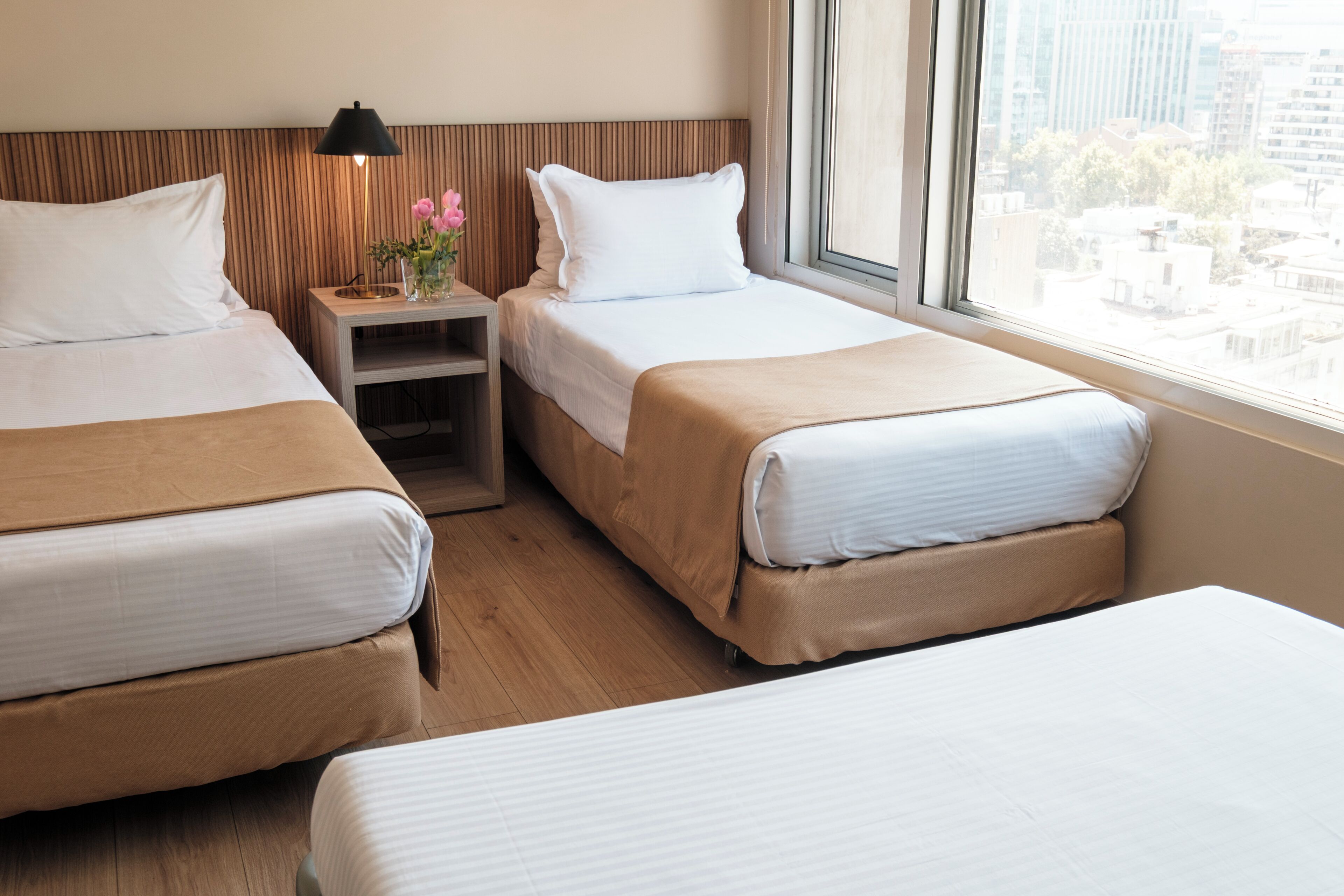 Habitación de hotel con dos camas, una mesita de noche con lámpara y ventana con vista a la ciudad.
