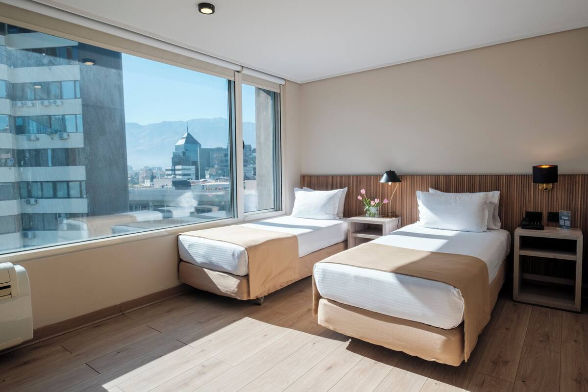 Una habitación de hotel con dos camas, un ventanal amplio con vistas a la ciudad, suelo de madera y una mesita de noche con lámpara.