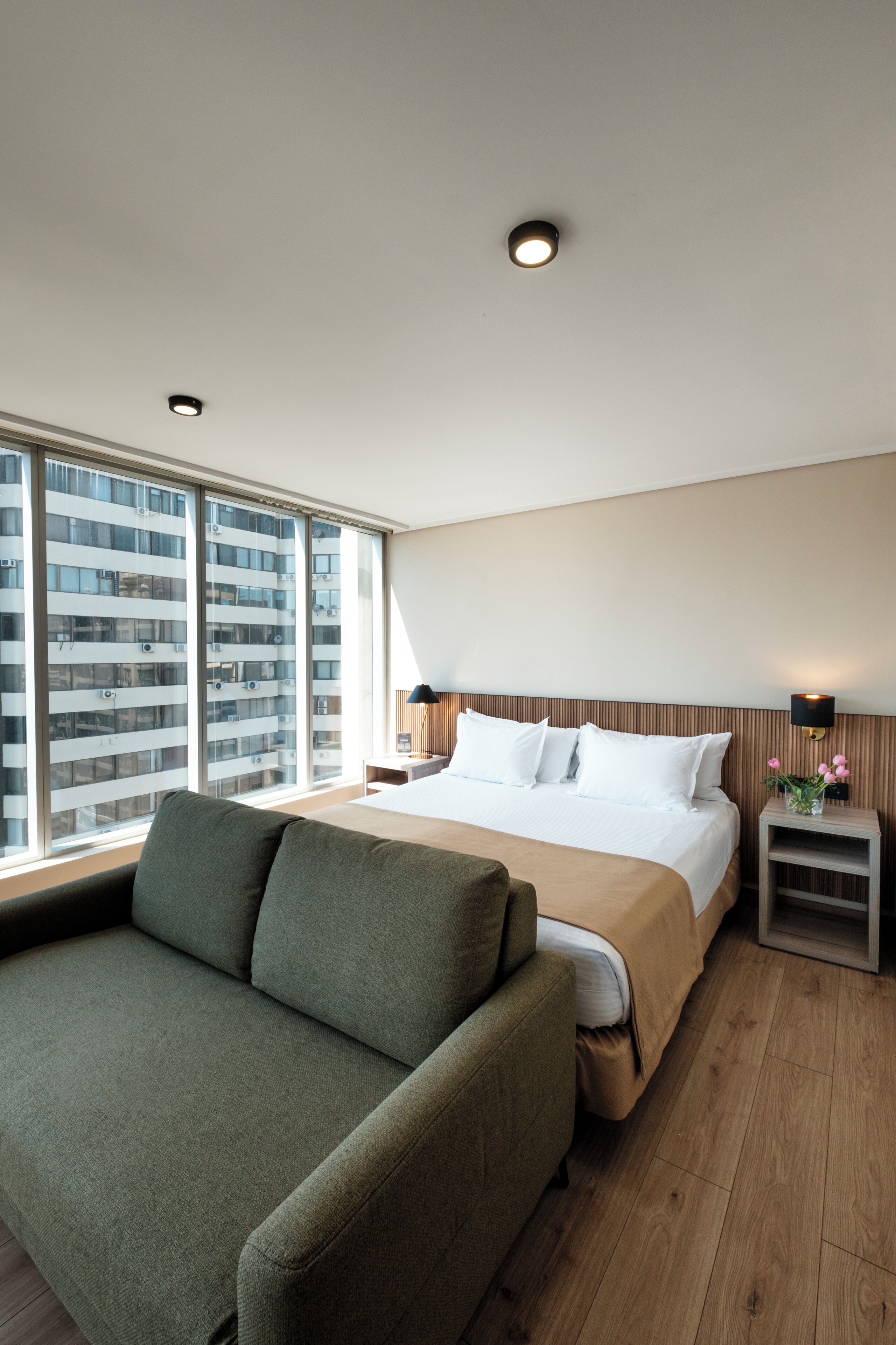 Habitación de hotel con cama, sofá, mesita de noche con lámpara y vista a un edificio a través de amplias ventanas.