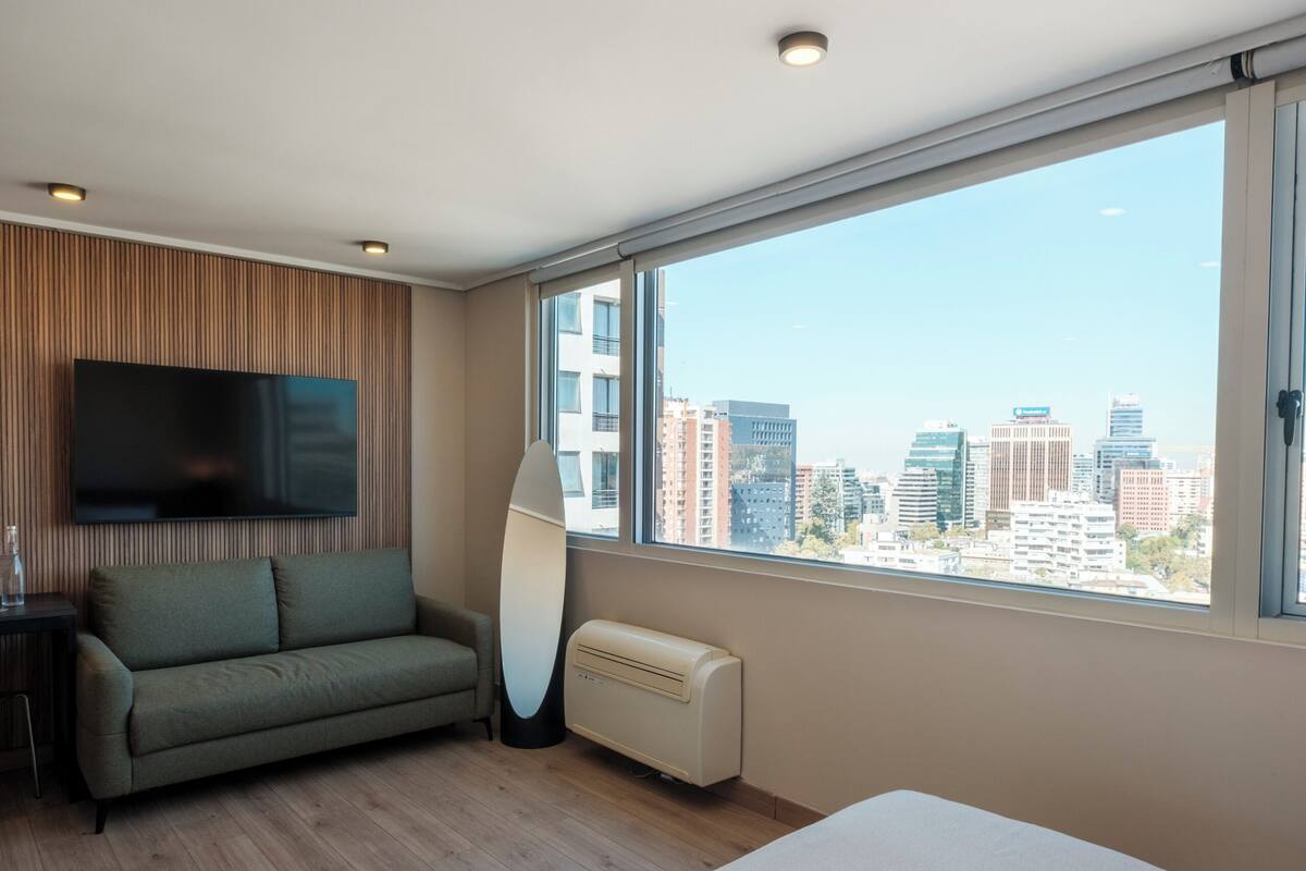 Una habitación de hotel moderna con cama, sofá, televisión de pantalla plana, un ventanal grande con vista a la ciudad y un radiador.