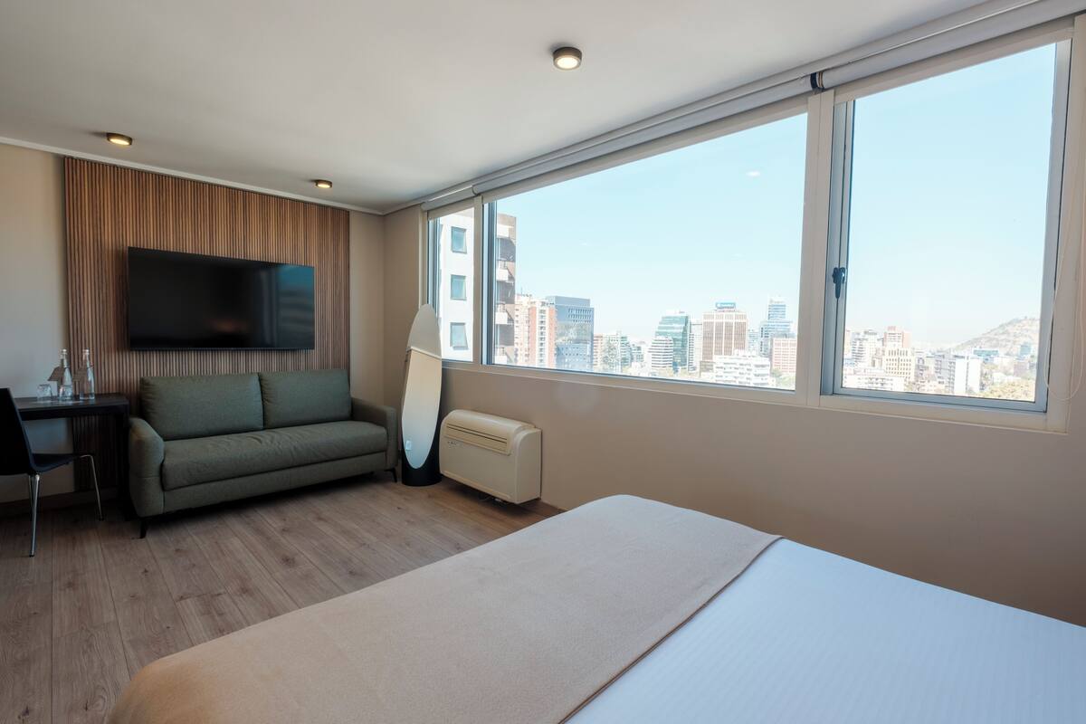 Una habitación de hotel moderna con cama, sofá, escritorio, televisor de pantalla plana y un amplio ventanal con vistas a la ciudad.
