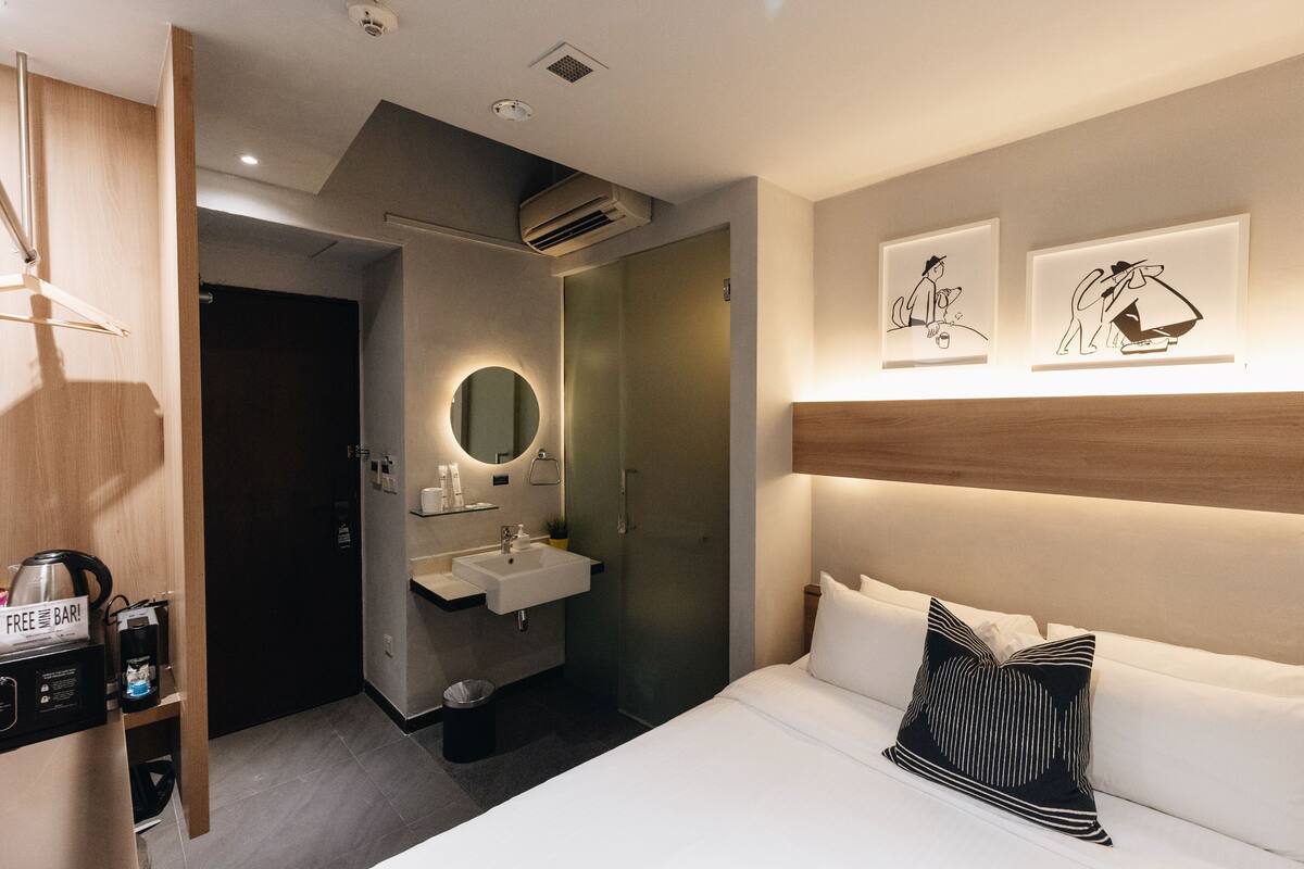 A modern hotel room with a bed, a bedside table, a wall-mounted shelf, a bathroom with a sink and mirror, and framed artwork on the wall.