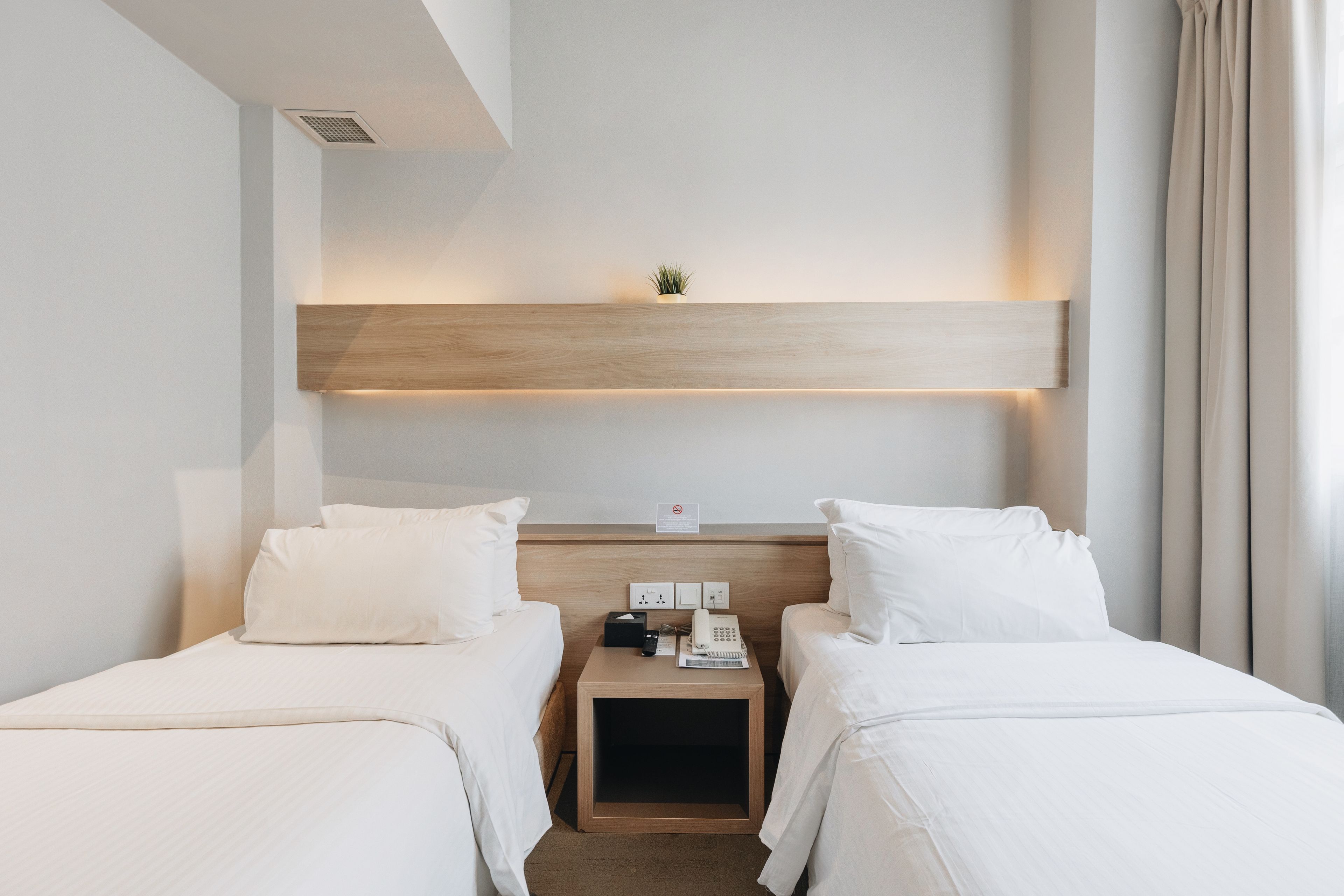A hotel room with two beds, a bedside table with a phone and a lamp, a small plant on the wall, and a window with curtains.