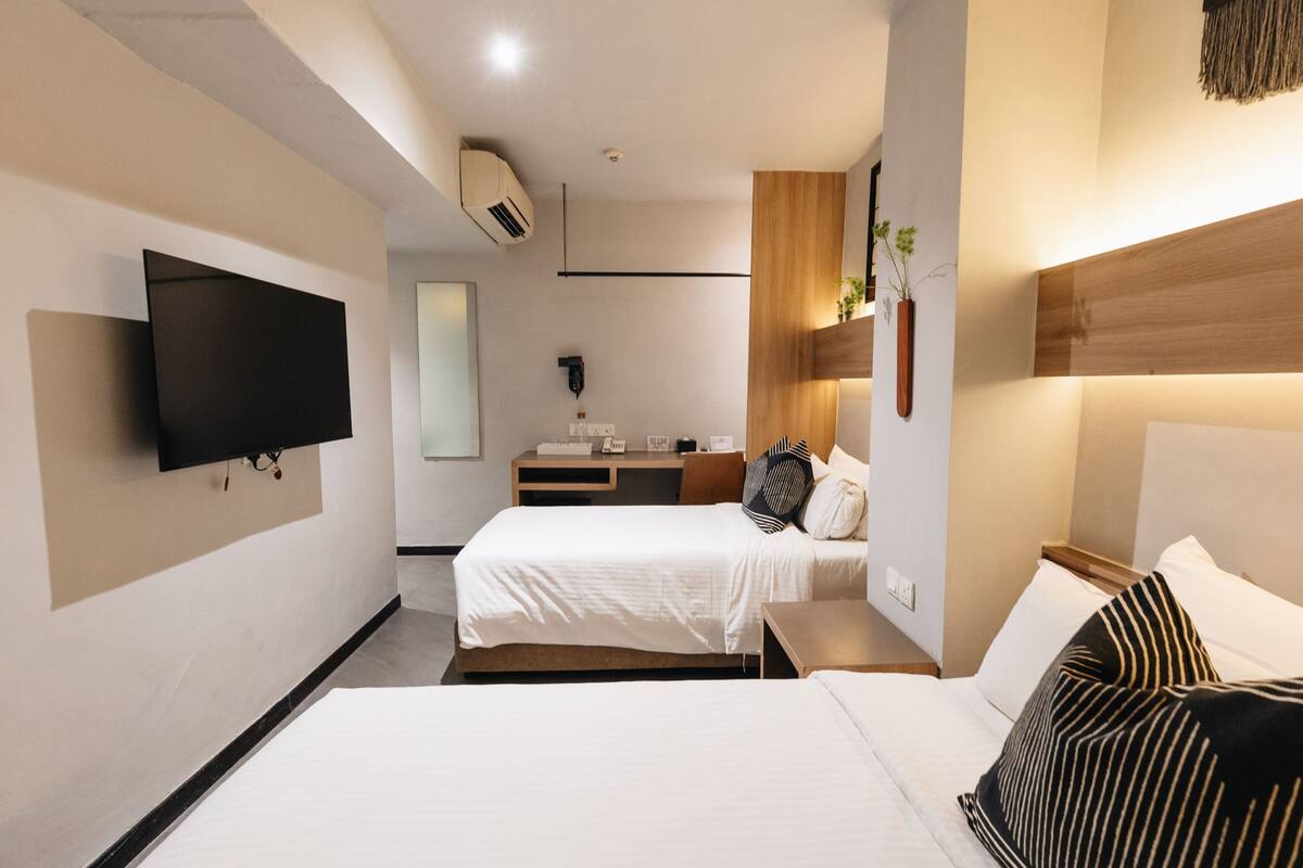 A modern hotel room with a large flat-screen TV mounted on the wall, a bed with white bedding, a bedside table, and a wooden headboard.