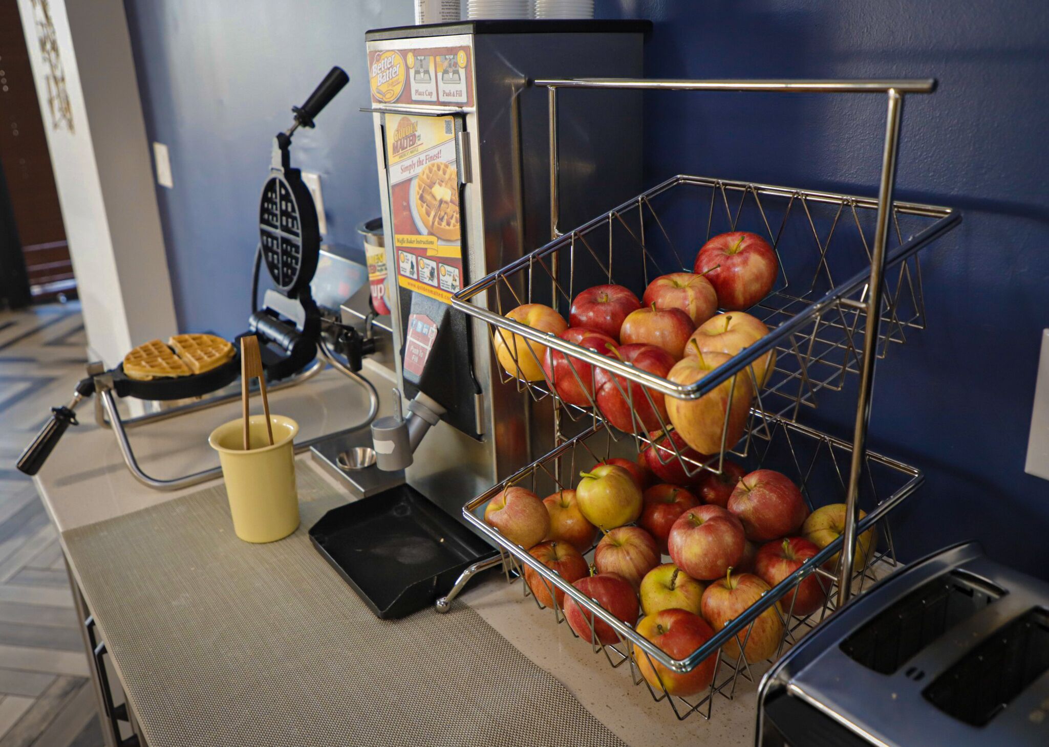 Un coin petit-déjeuner équipé d’un appareil à gaufres, d’une machine à café et d’un panier de fruits contenant des pommes.