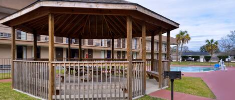 A wooden gazebo with a sloped roof, surrounded by a wooden fence, in front of a building with balconies and a swimming pool.