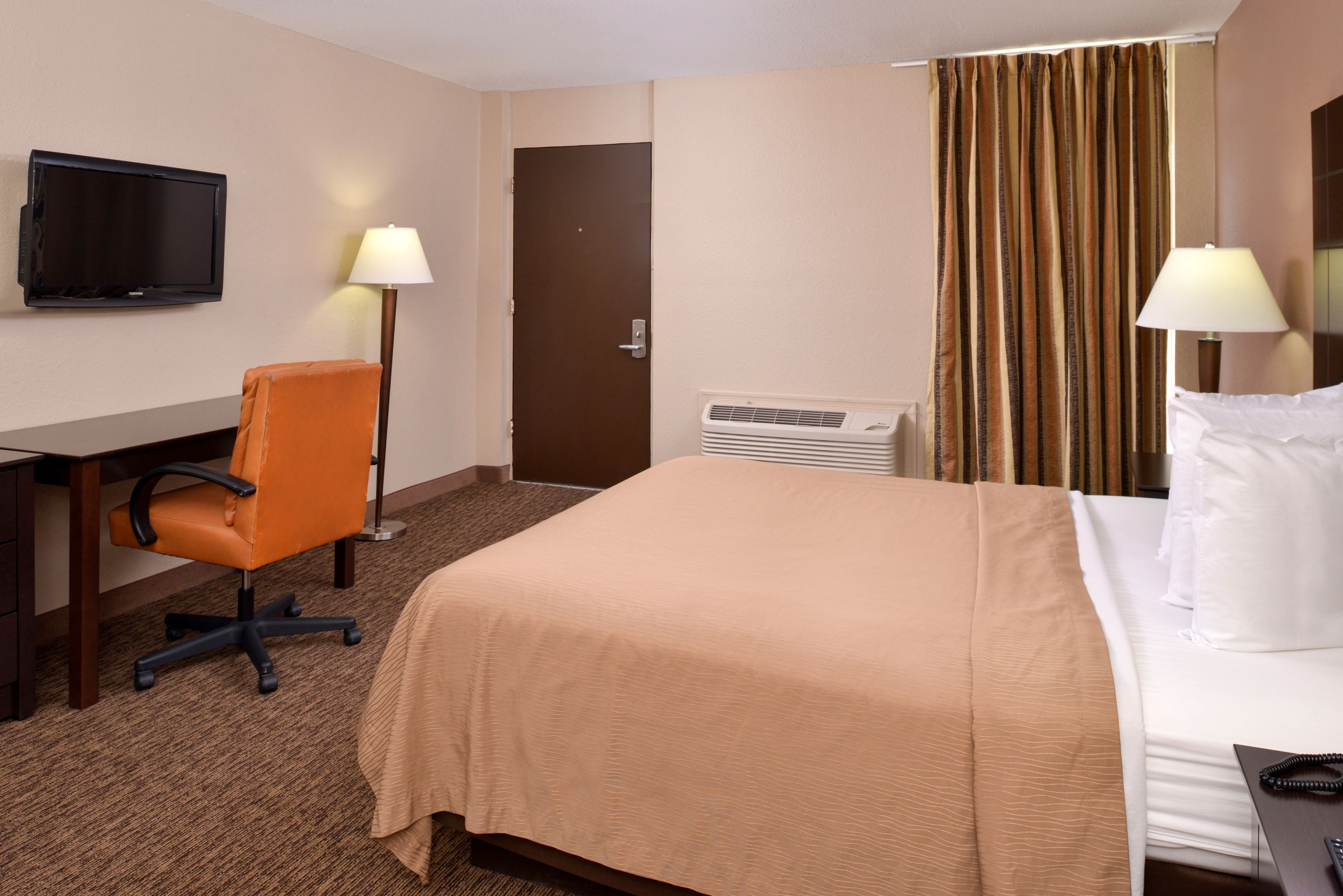 Habitación de hotel con cama, escritorio, silla, televisor, aire acondicionado y cortinas.