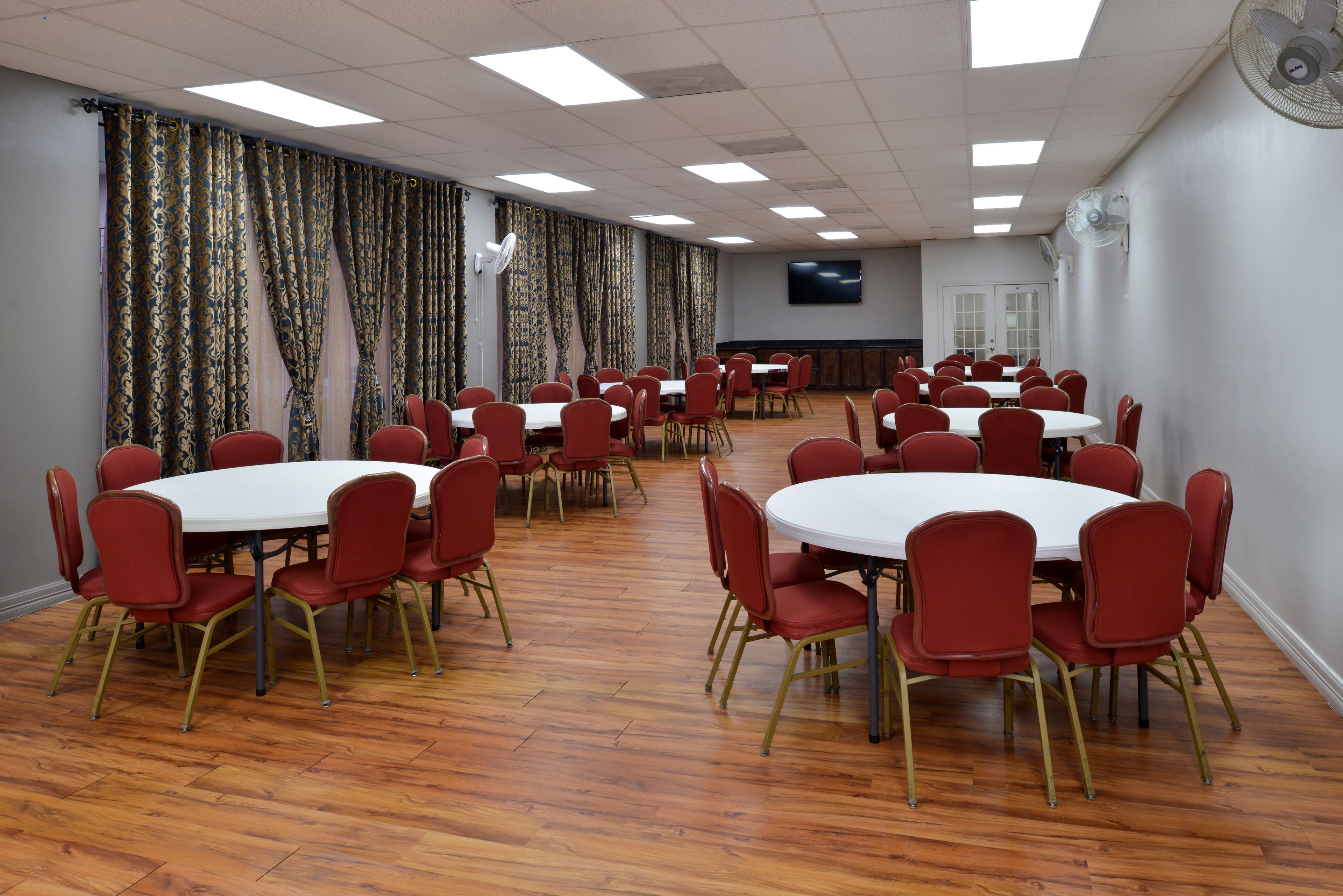 A conference room with multiple round tables and red chairs, equipped with ceiling fans and a flat-screen TV.