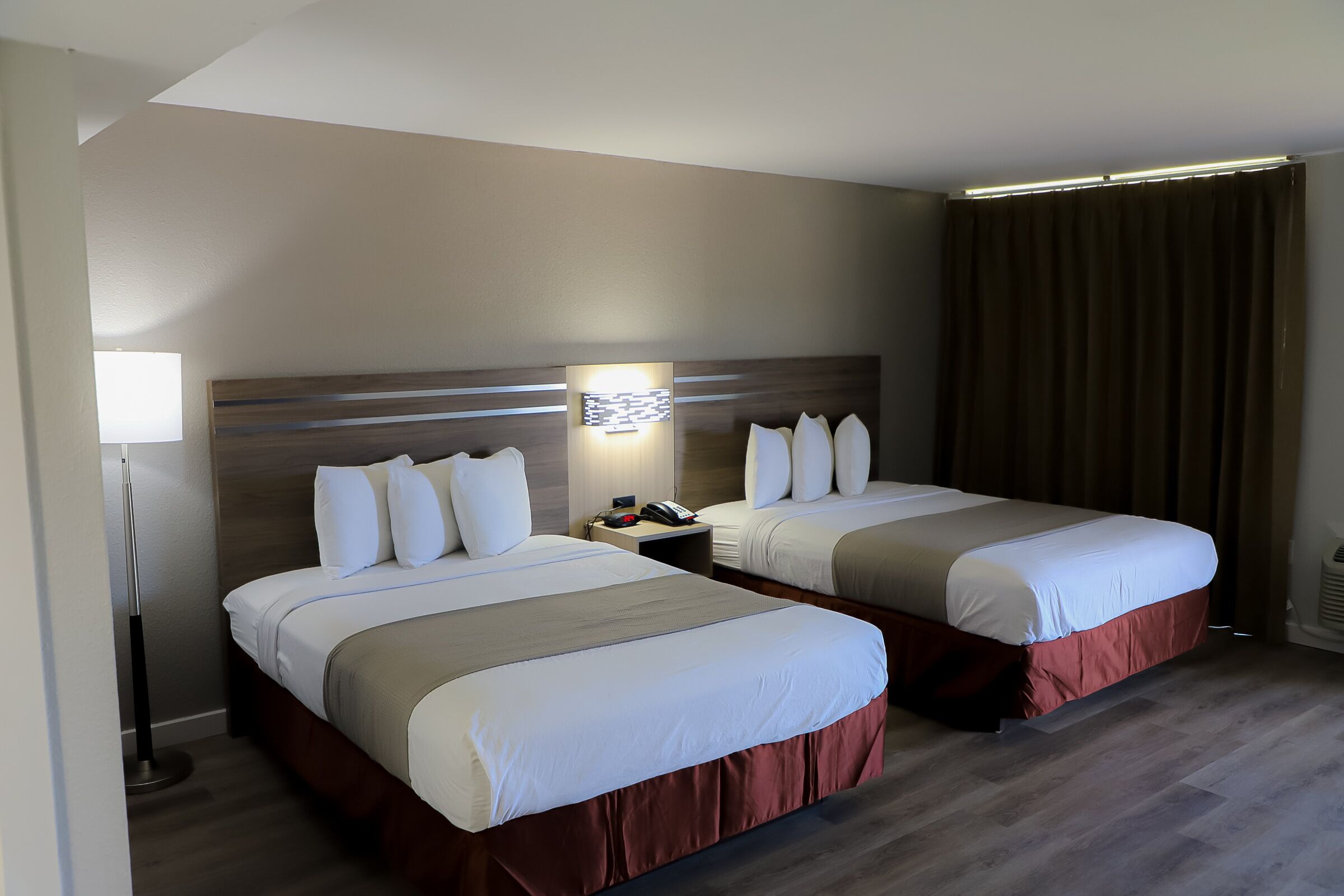 Hotellihuone, jossa on kaksi sänkyä, yöpöytä, lamppu ja ikkuna, jossa on verhot.