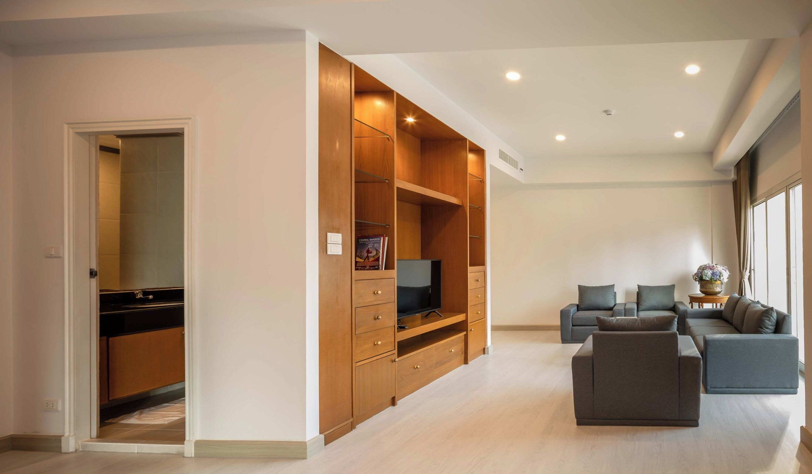A modern living room with a grey sofa set, a wooden entertainment unit with a TV, and a large window providing natural light.