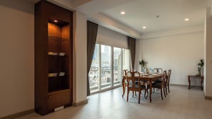 A dining area with a round table and chairs, a wooden cabinet, and a large window with a view of buildings. - Chaidee Mansion (Bangkok)