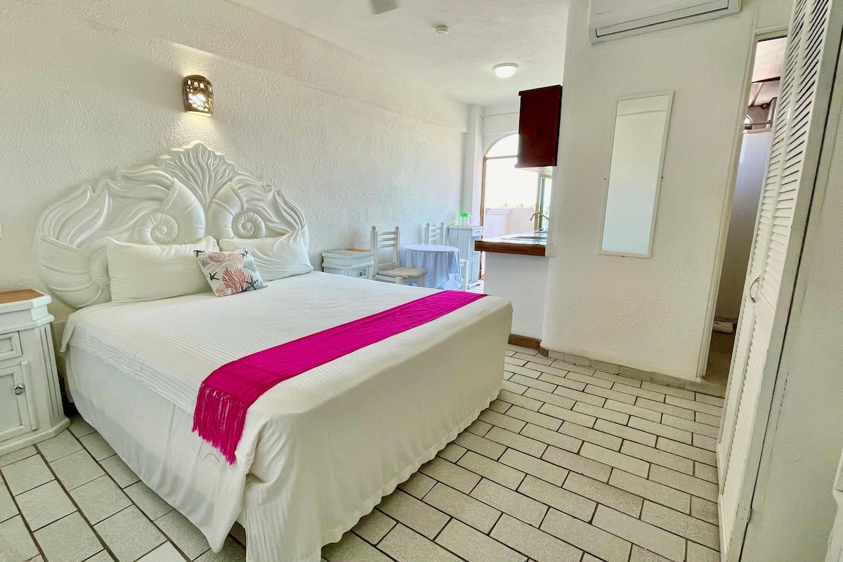Un dormitorio con una cama grande, una mesita de noche, un aparador, una ventana con persianas y una puerta que da a otra habitación.