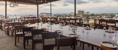 An outdoor dining area with multiple tables and chairs, overlooking a cityscape and the ocean.