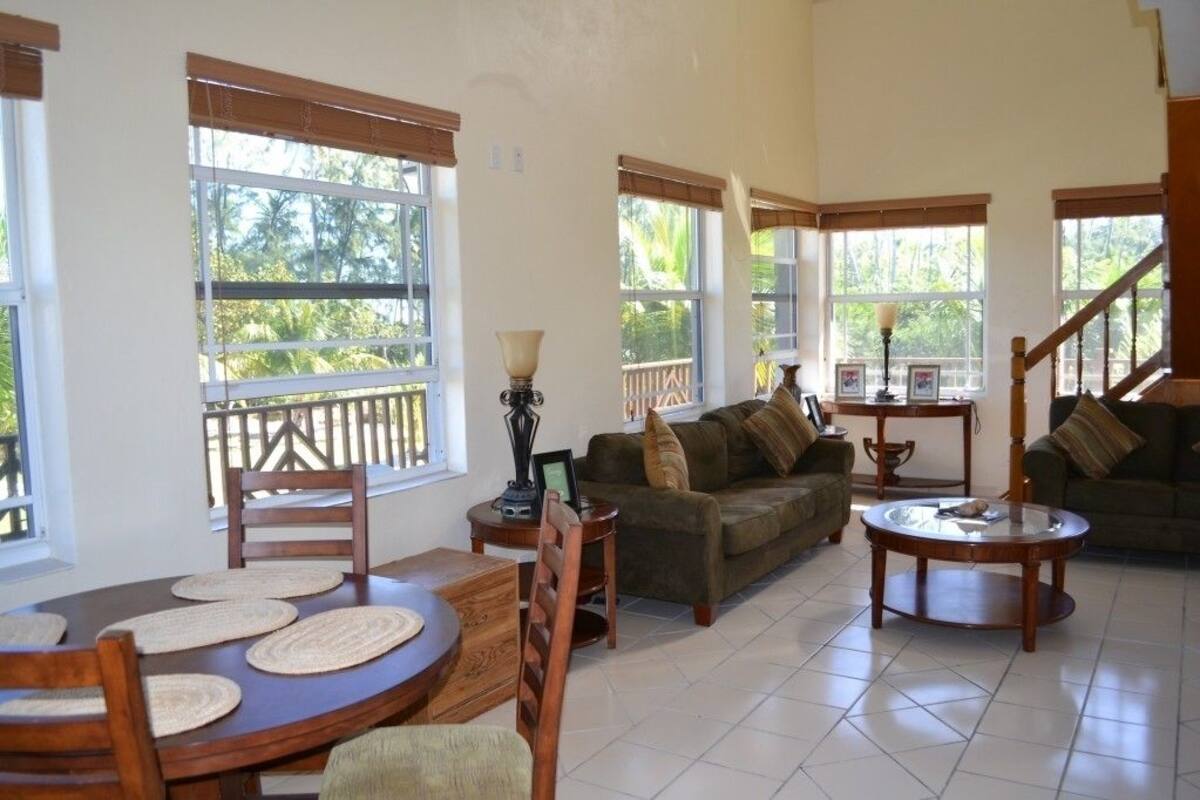 A spacious living area with a dining table, two sofas, and a coffee table. Large windows provide natural light and views of greenery outside.