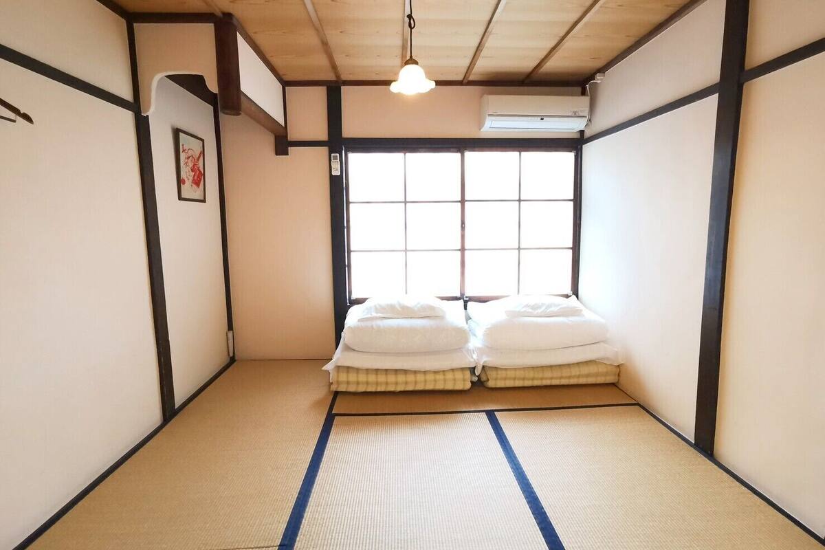 A traditional Japanese room with tatami flooring, a futon bed, a window with a view, and an air conditioning unit.