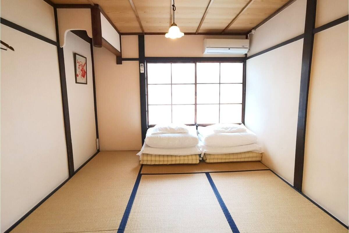 A traditional Japanese room with tatami flooring, a futon bed, a window with a grid pattern, and a hanging light fixture.