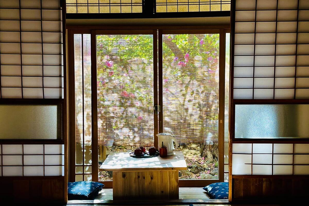 A traditional Japanese room with tatami flooring, a low wooden table with tea set, and a view through shoji screens to a garden.